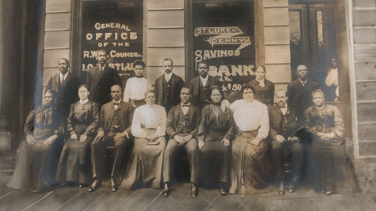 A sepia-toned photograph shows black people dressed in clothes from around 1900 seated outside a bank