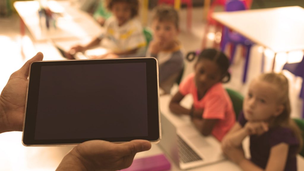 An adult holds a tablet in the foreground with a row of schoolchildren in the background