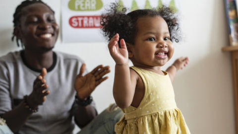 A toddler in a yellow dress while her dad claps in the background