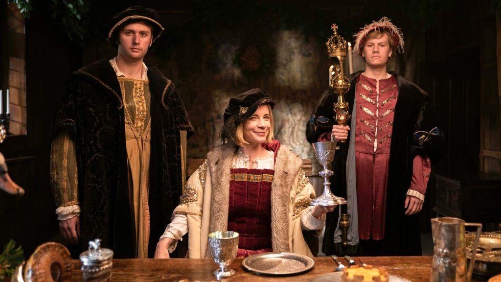 Lucy Worsley, dressed in Tudor garb, sits at a table with two men, also in costume, standing on either side