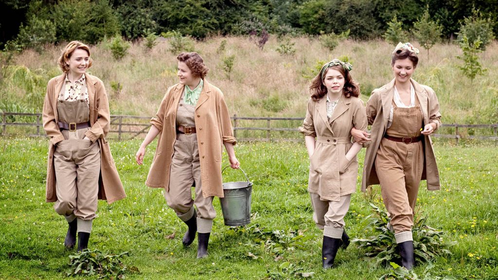 Four women in tan farm clothes walk through a grassy area