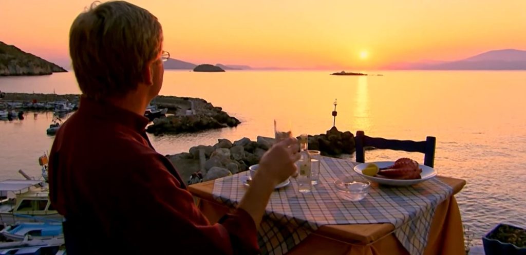Rick Steves holding a drink at a table viewing an ocean and sunset