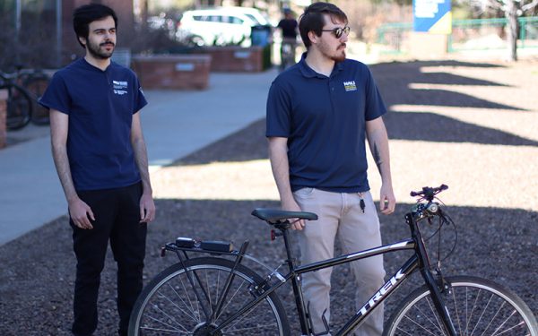 Two men wearing NAU shirts stand next to a bike on a sidewalk