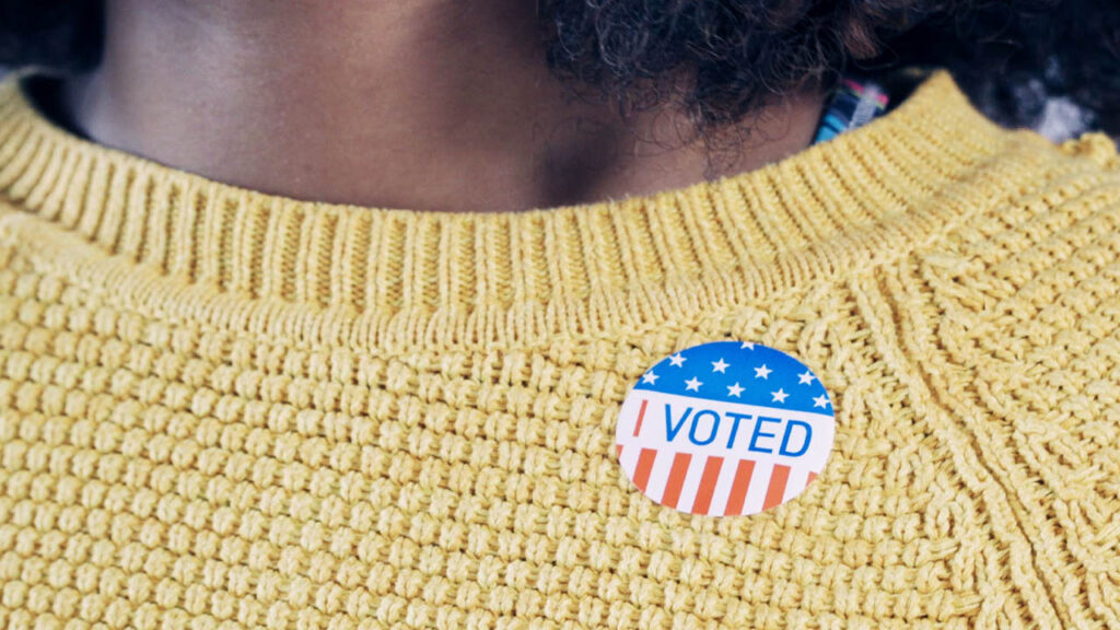 A Black person with natural hair wears an I Voted sticker on a yellow sweater