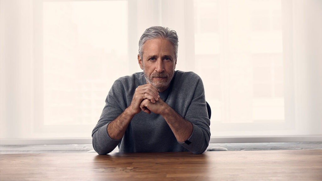Jon Stewart sits at a table with his head held up by his hands