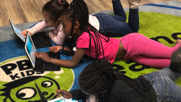 Three young children lie on a PBS KIDS rug as they play games on tablets