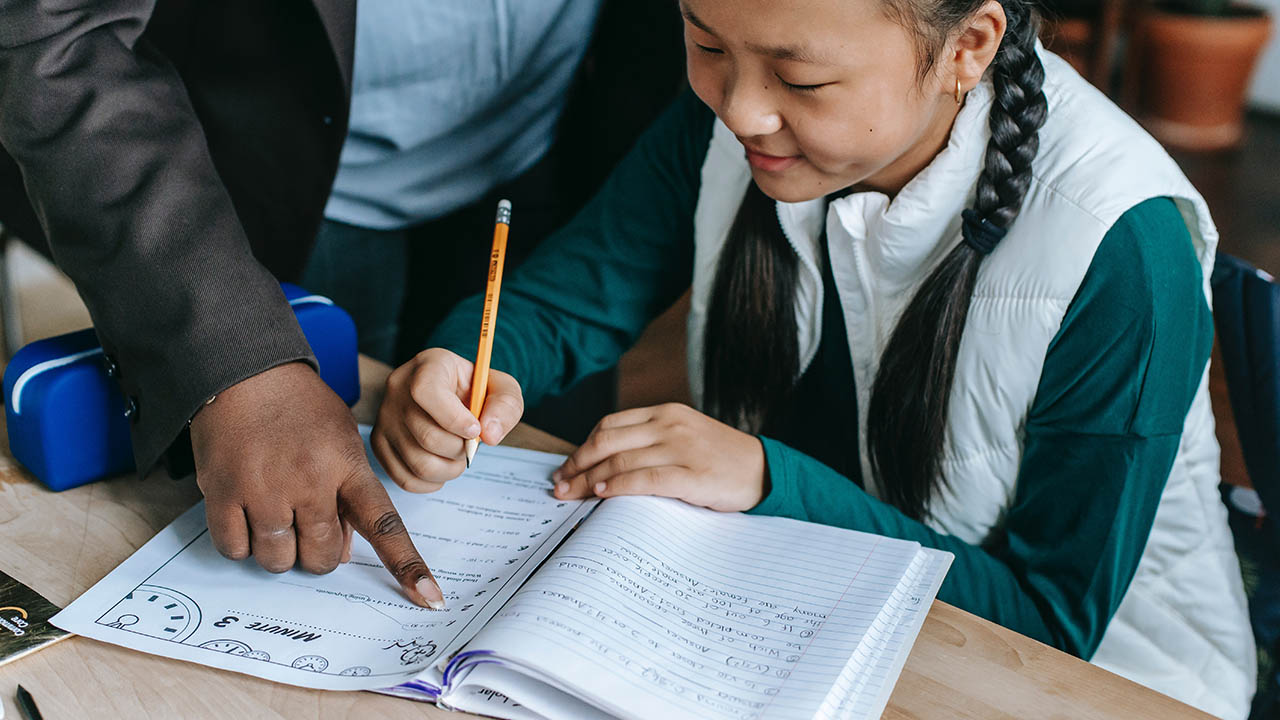 A Black teacher helps a girl with a worksheet.