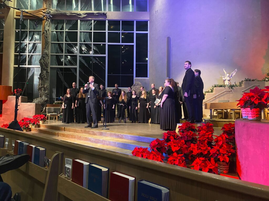 Phoenix Chorale standing on stage at Pinnacle Presbyterian church. Chris Gabbitas is speaking to the audience with a microphone. The stage is decorated with red poinsettias 