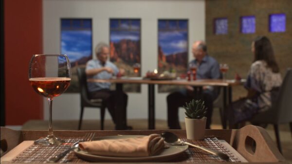 A place setting with a wine glass are set on a tray just in front of the camera, with three people sitting around a table in the background.