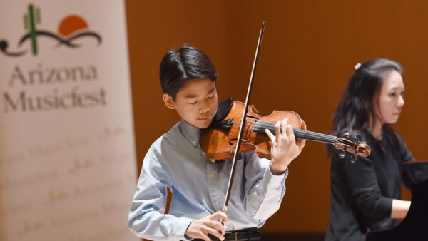 young Asian boy playing violin. He is wearing a blue button down shirt. A woman is playing piano in the background. There is also a banner in the background that says 
