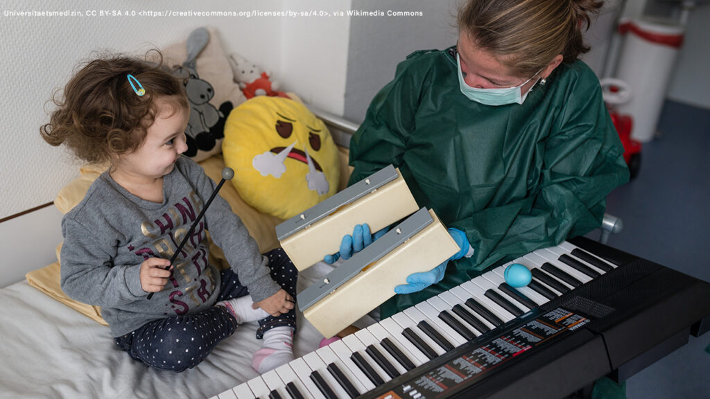 woman wearing a mask and a green surgical gown sits on a hospital bed holding xylophone blocks. A toddler girl sits next to her holding a beater. There is a keyboard in front of them on the bed. There is a yellow emoji pillow behind them.
