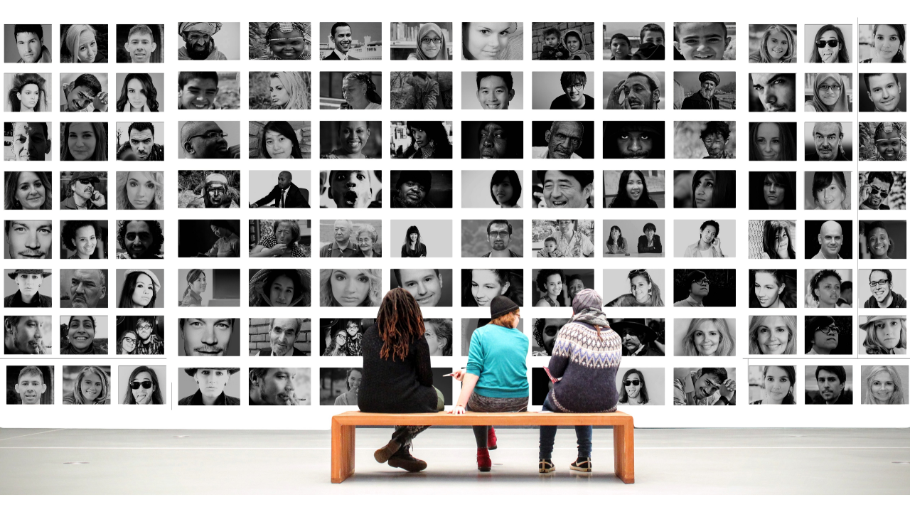 3 people sitting on a bench in front of a photo wall. The photos are black and white and are all of faces of people of all different ages and races