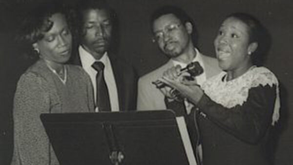 An African American woman sings from a stage as part of a quartet