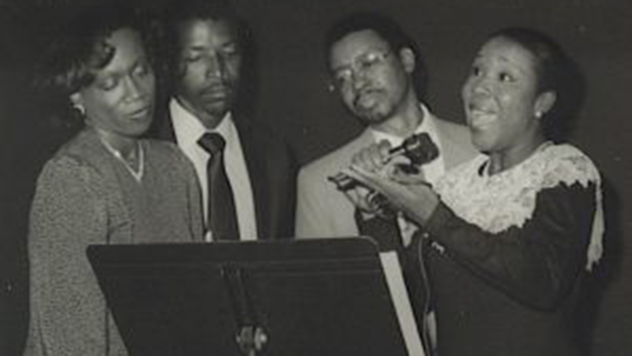 An African American woman sings from a stage as part of a quartet