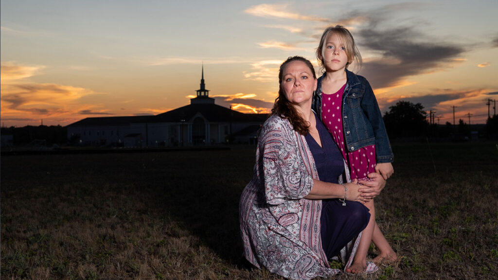 A woman kneels and holds her daughter to her as they stand in front of a sunset.