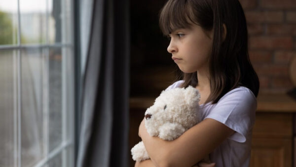 A young girl holds a stuffed animal and looks out the window sadly.
