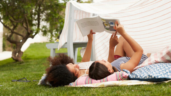 A woman and her child read a book while laying under a blanket fort outside.