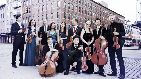 11 members of the Manhattan Chamber Players holding string instruments standing outside in front of a large brick building.