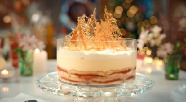 A trifle sits in a dish on a clear, crystal serving stand on a table set for the holiday season