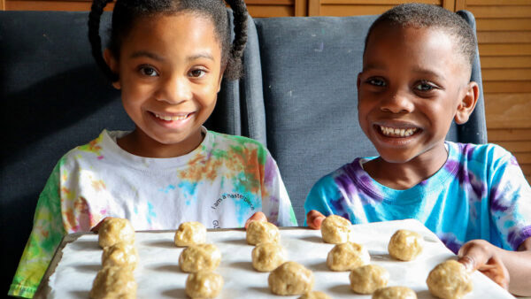 Two young children smile into the camera holding a cooking sheet with balls of cookie dough on it