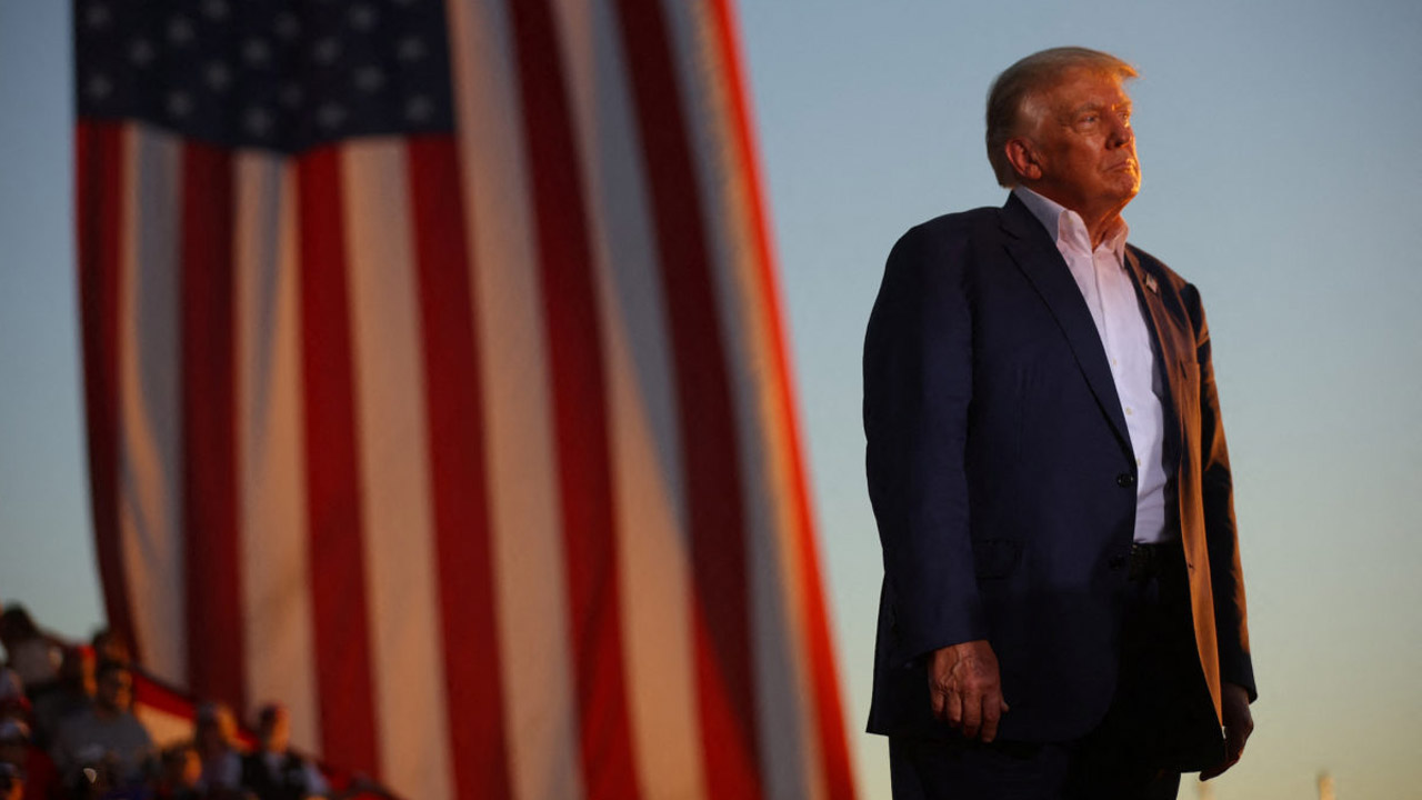 Donald Trump standing on a stage with a huge American Flag behind him