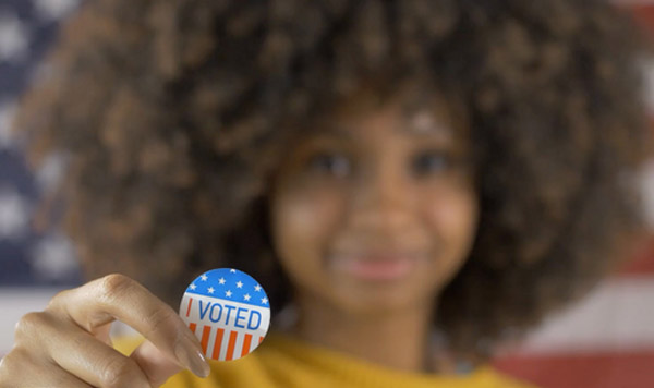 A young girl holds a button that reads: Vote