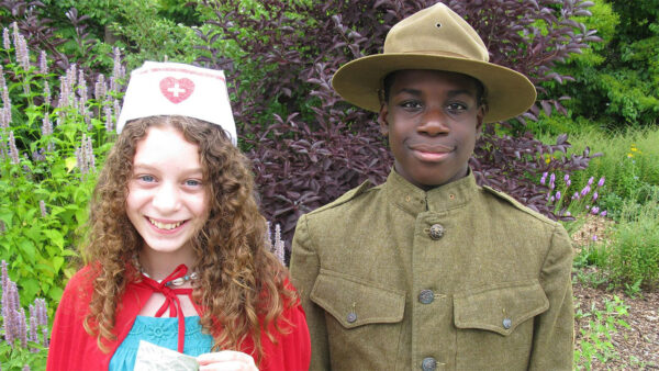 A young girl dressed as a nurse stands beside a boy in a soldier uniform, both smiling and posing for the camera.