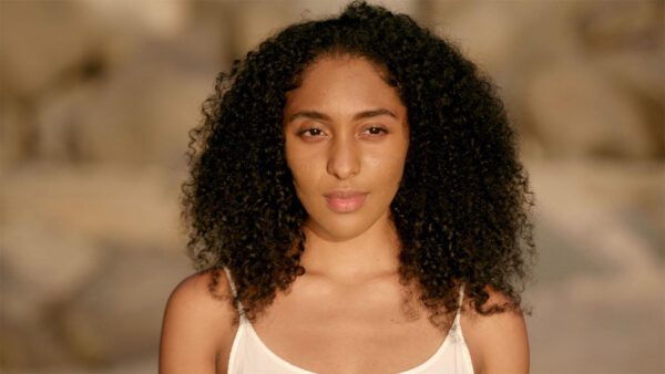 A young woman with curly hair wearing a white tank top, smiling and standing against a neutral background.