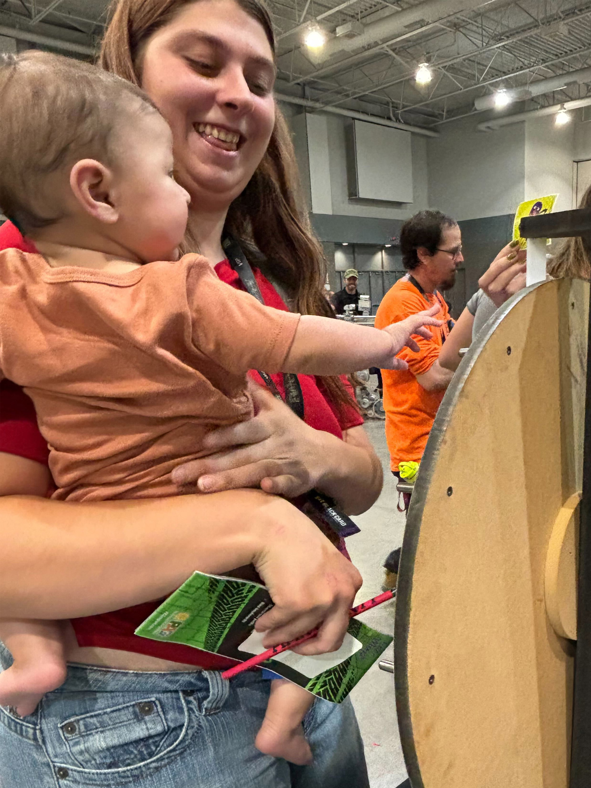 An adult holding a child interacts with a circular machine, it appears to be an indoor event.