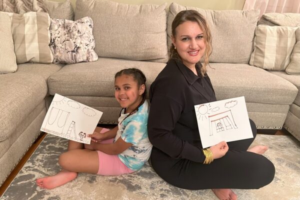 A mom and daughter sit back to back holding drawings