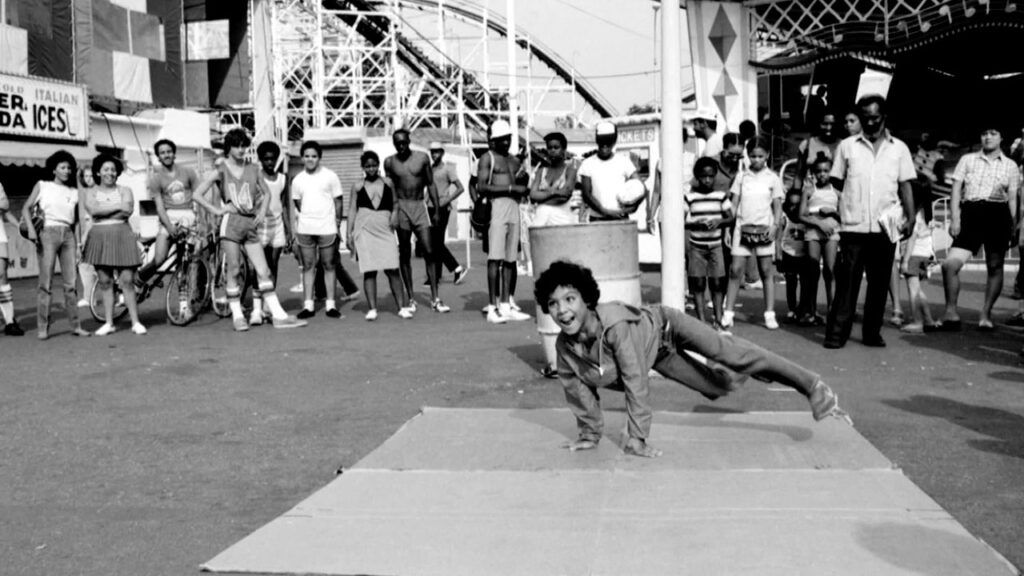 A woman performs a handstand on a mat outside an amusement park, showcasing her skills in front of an engaged crowd.