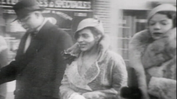 A vintage black and white scene from the 1930s featuring a woman and man strolling together down a city street.