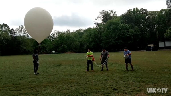 Three individuals stand in a grassy field, holding a large white balloon against a clear blue sky.