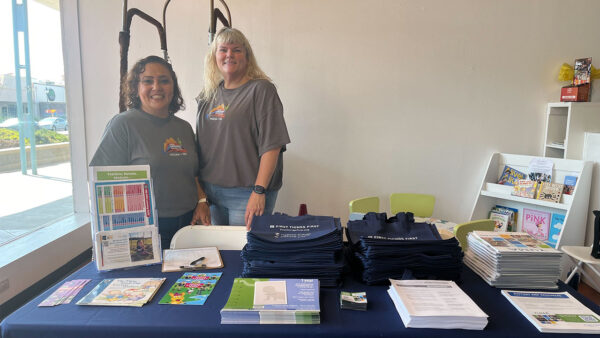 Two women stand behind a table with booklets at an event or resource fair.