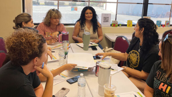 A group of women seated at a table, engaged in discussion while reviewing papers and documents.