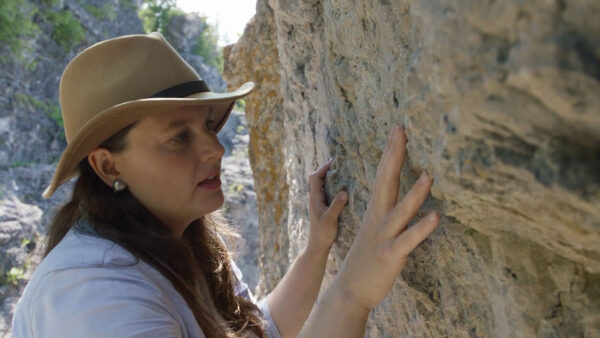 A woman wearing a hat gently touches a rock while standing outdoors.