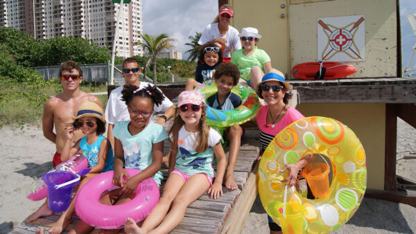 A group of kids smiling and posing together for a photo on a sunny beach with waves in the background.