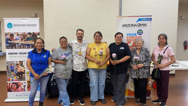 A diverse group of people stands together in front of a large banner of AZPBS
