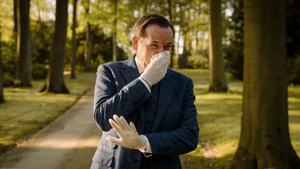 A man dressed in a suit and gloves is blowing his nose with a tissue.