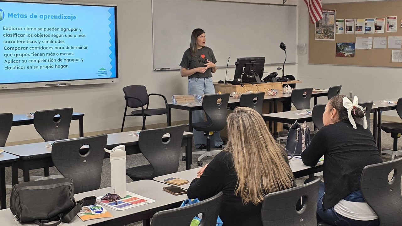 A women instructs students sitting at desks in a classroom setting.