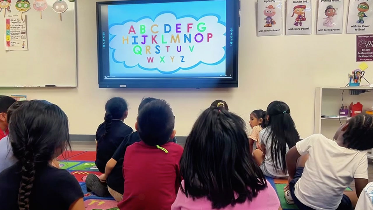 Children in a classroom watching a teacher's lesson displayed on a screen.