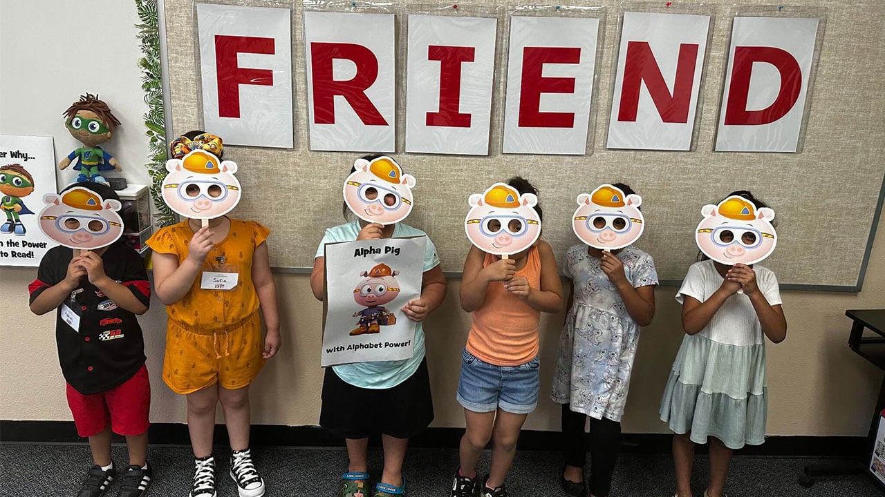A group of children holding their masks in front of a sign that reads "Friend."