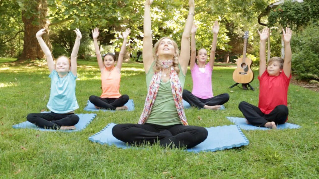 Children practicing yoga poses on mats in a sunny park, surrounded by trees and grass.