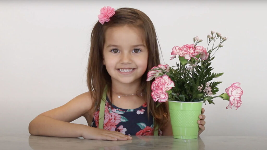 A little girl smiles while holding a flower in a green vase.