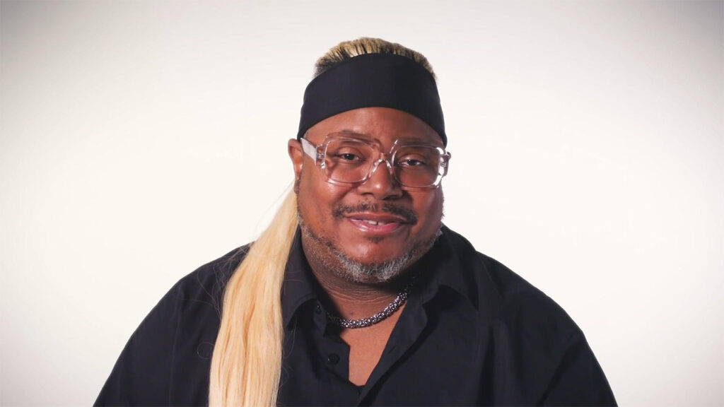 A man wearing glasses and a long blonde wig, smiling at the camera.