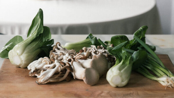 A wooden cutting board topped with sliced mushrooms alongside various green vegetables