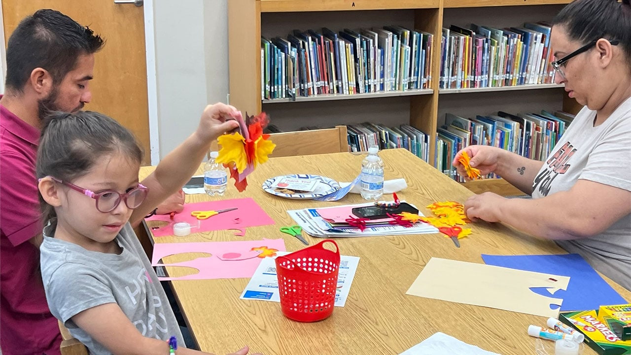 A family of three sits at a table, engaged in crafting with paper and scissors
