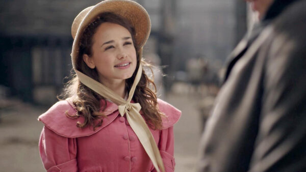 A woman in a pink dress and hat engages in conversation with a man outdoors.