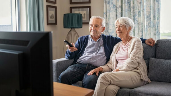 An older couple sits on a couch watching television