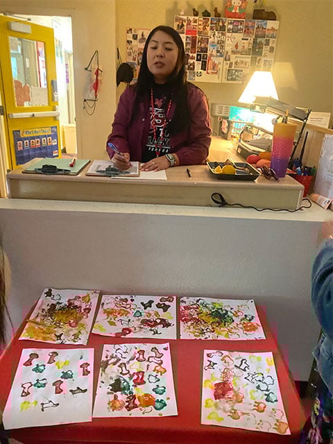 A woman sits at a desk surrounded by various art supplies and creations, focused on her artistic work.